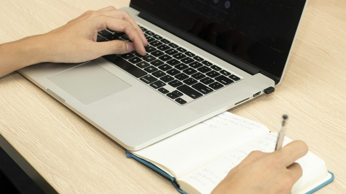 Mâini care lucrează la laptop și notează într-un caiet, ca să verifice că asistentul AI răspunde corect clienților