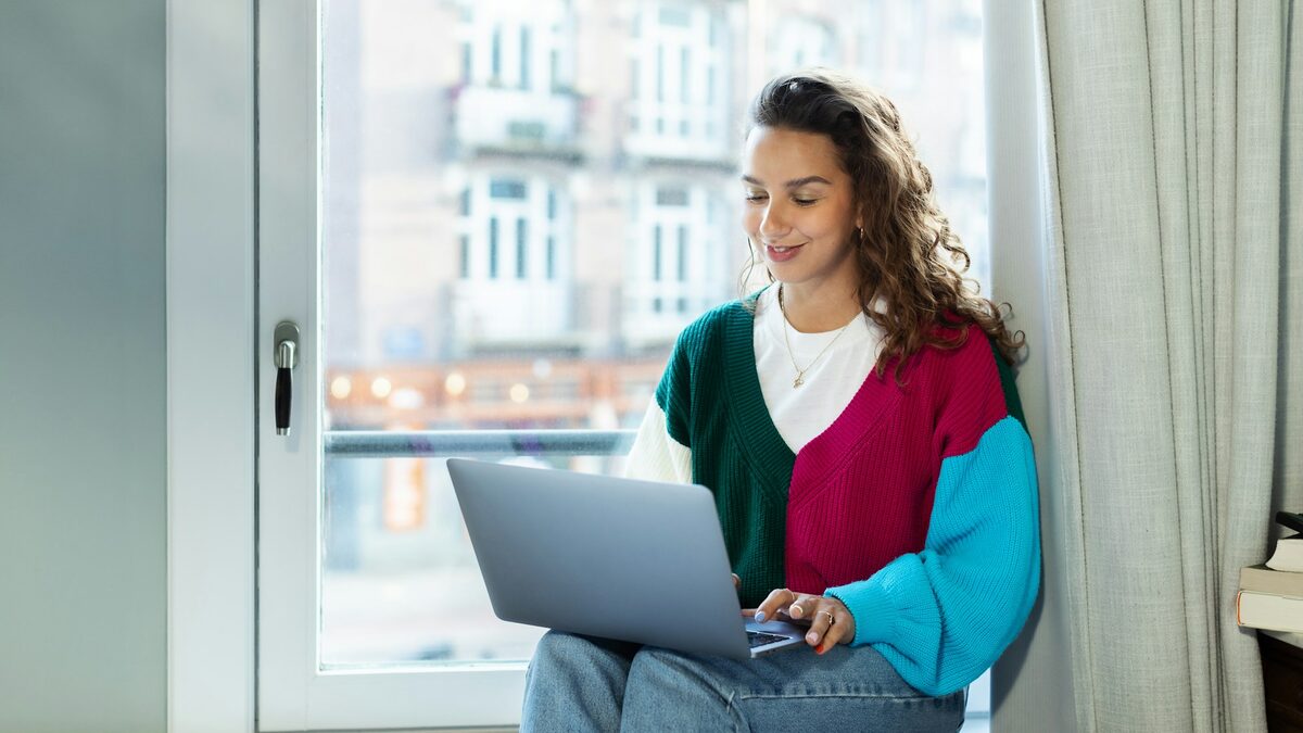 Tânără freelanceră la fereastră, cu laptopul pe genunchi, care își scrie profilul pe platformele de freelancing
