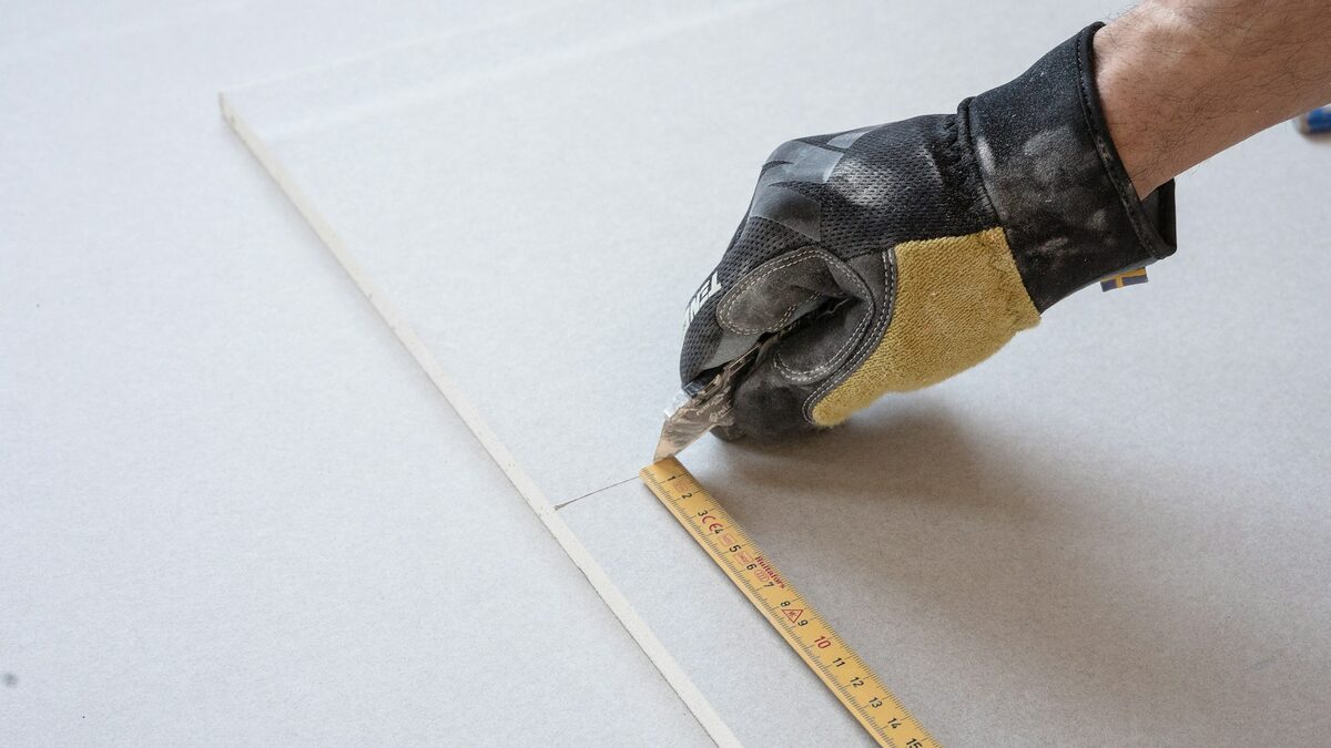 Mână de meseriaș cu mănușă care măsoară o placă de rigips cu metrul, pentru calculul materialelor