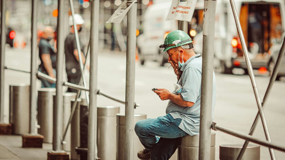 Meseriaș cu cască de protecție care completează pe telefon devizul pentru o lucrare, într-o pauză pe șantier