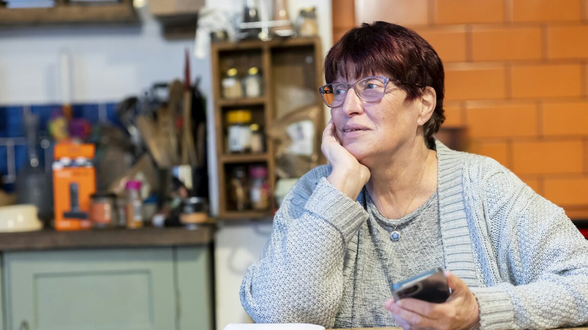 Bunică în bucătărie, cu telefonul în mână, pregătită să folosească asistentul vocal ca să-și sune nepoata