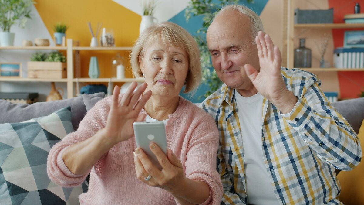Doi bunici pe canapea care salută într-un apel video pe telefon, de Ziua Internațională a Vârstnicilor