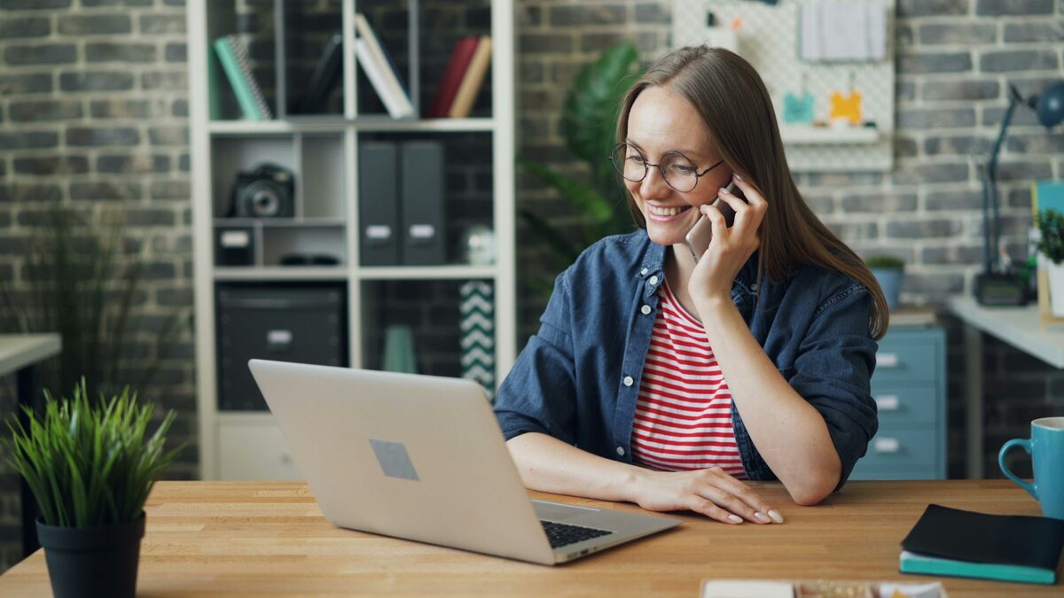 Femeie zâmbitoare care vorbește la telefon în fața laptopului, ca atunci când testezi un agent vocal AI înainte să îl alegi