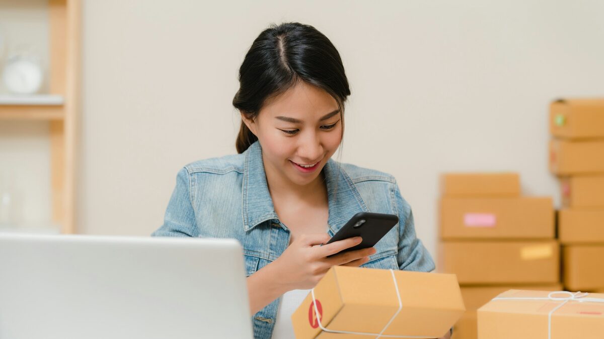 Antreprenoare care fotografiază cu telefonul un colet, lângă laptop, pentru catalogul de produse pentru Google Shopping și eMAG