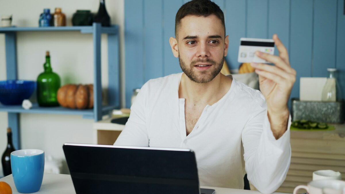 Bărbat în fața laptopului, cu cardul în mână, care face compararea prețurilor înainte să plătească o comandă online