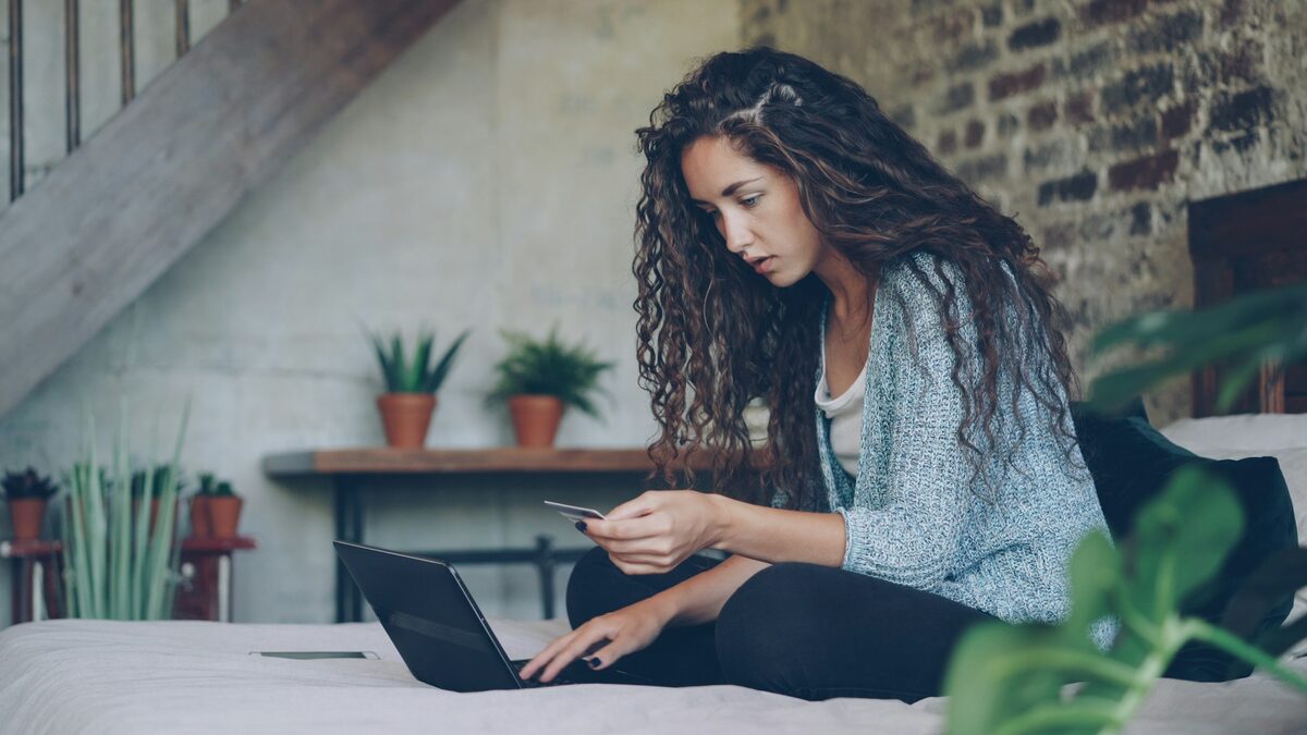 Femeie cu cardul în mână în fața laptopului, care se gândește înainte să finalizeze comanda, evitând cumpărăturile impulsive