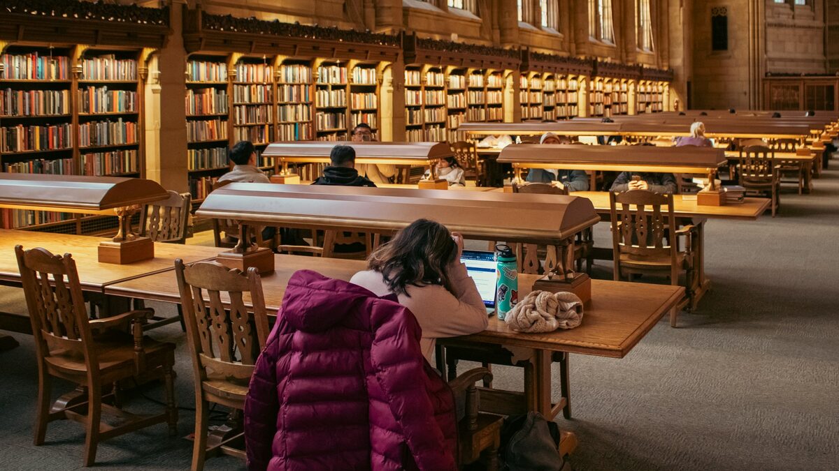 Studentă care învață într-o sală mare de bibliotecă, printre rafturi cu cărți, și se întreabă de unde știe AI-ul ce știe