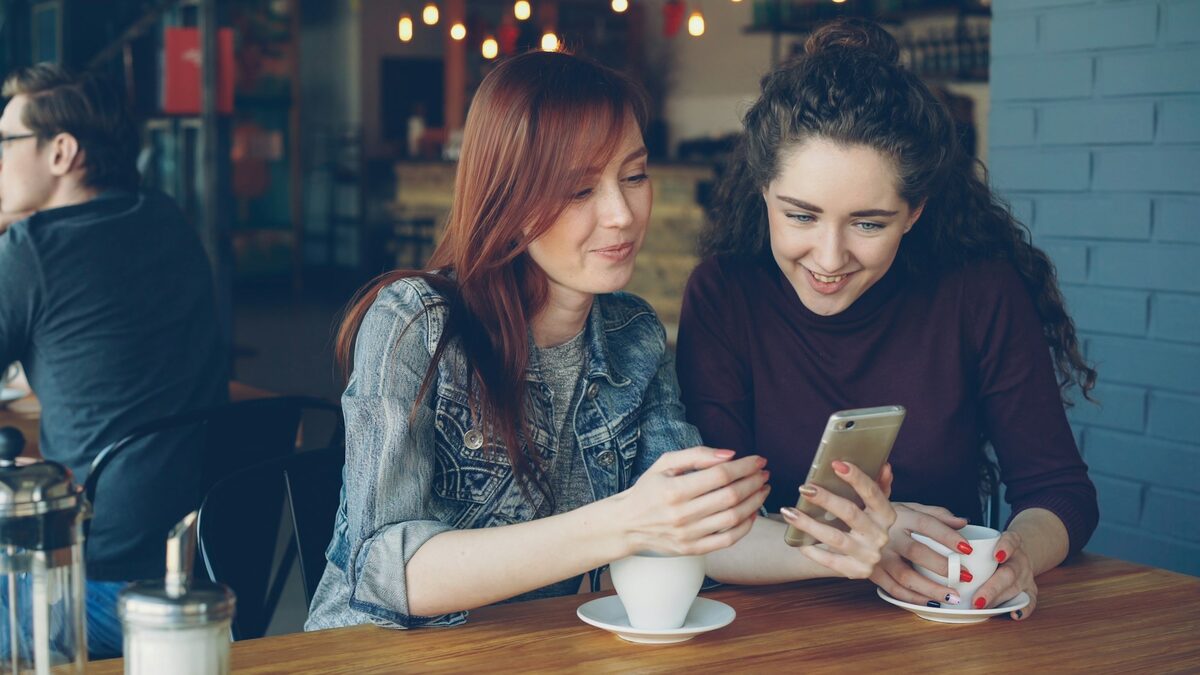 Două prietene la o cafenea care caută pe telefon o firmă, așa cum caută clienții în 2026