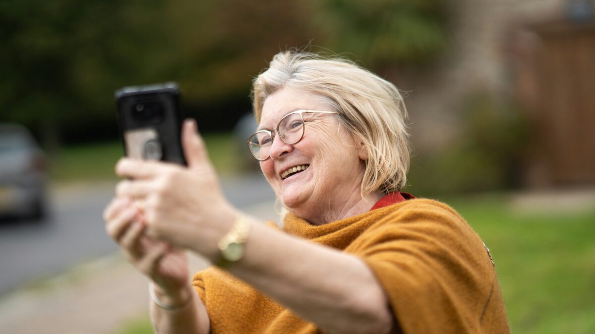 Bunică zâmbitoare cu telefonul în mână, care învață din emoji explicate pe înțeles