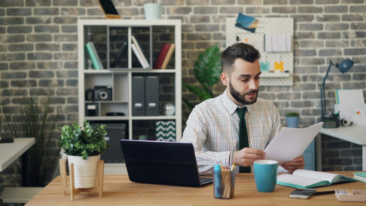 Bărbat care compară pe laptop un răspuns AI cu un document tipărit, întrebându-se dacă AI-ul știe tot