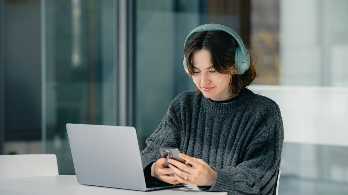 Tânără care își scrie scrisoarea de intenție pe laptop, cu anunțul de angajare deschis pe telefon
