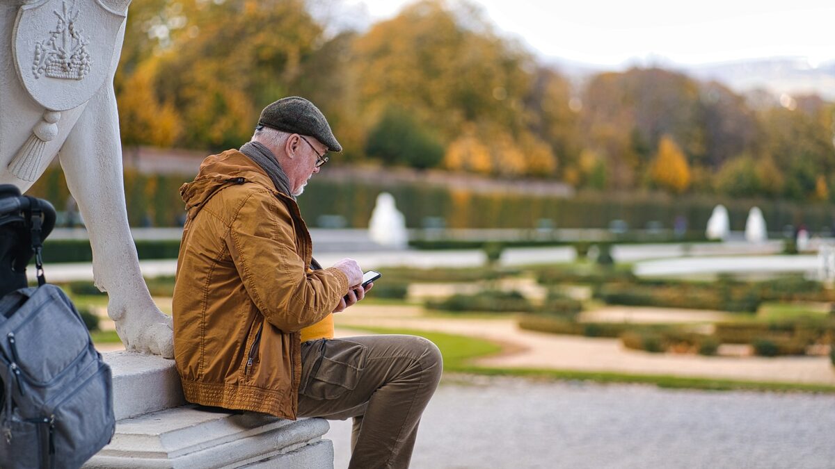 Bărbat în vârstă, pe o bancă în parc, care privește pe telefon o reclamă cu investiții false cu vedete