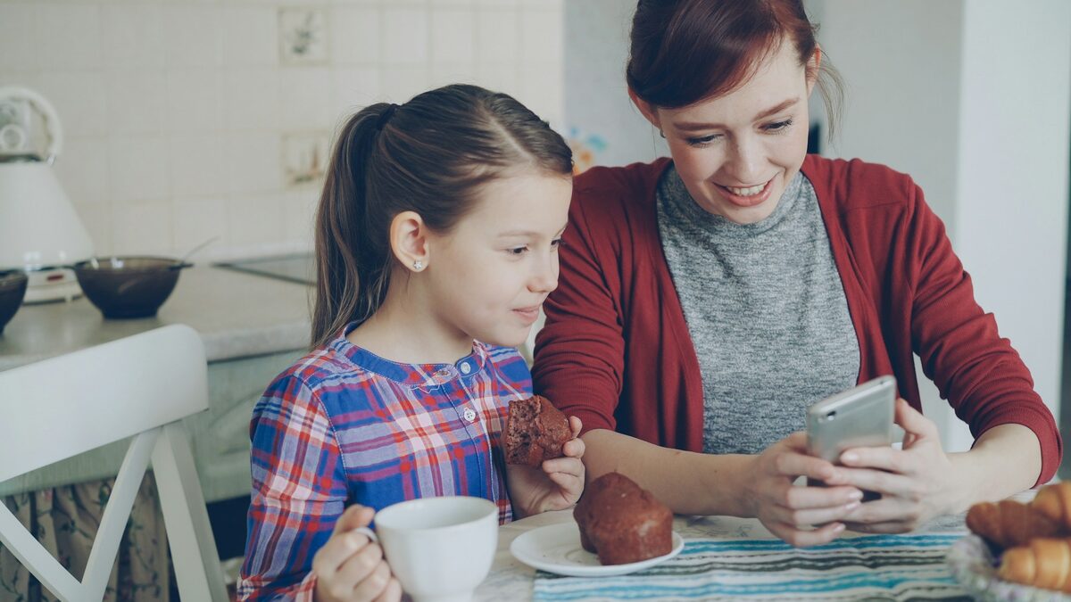 Mamă și fiică la masa din bucătărie, care stabilesc împreună pe telefon partajarea locației cu familia