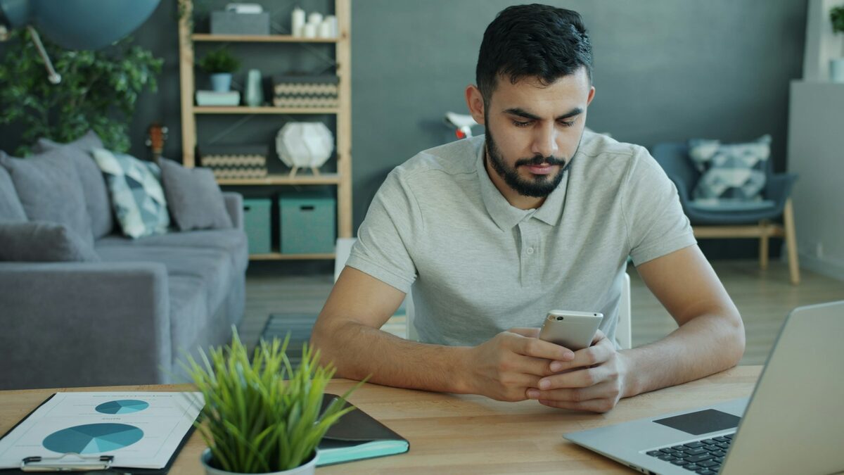 Bărbat la birou care face verificarea de securitate a contului Google, cu telefonul în mână pentru confirmare și laptopul alături