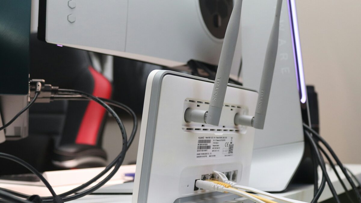 Router Wi-Fi alb cu antene, conectat prin cabluri pe birou; parola de Wi-Fi se află pe eticheta lui