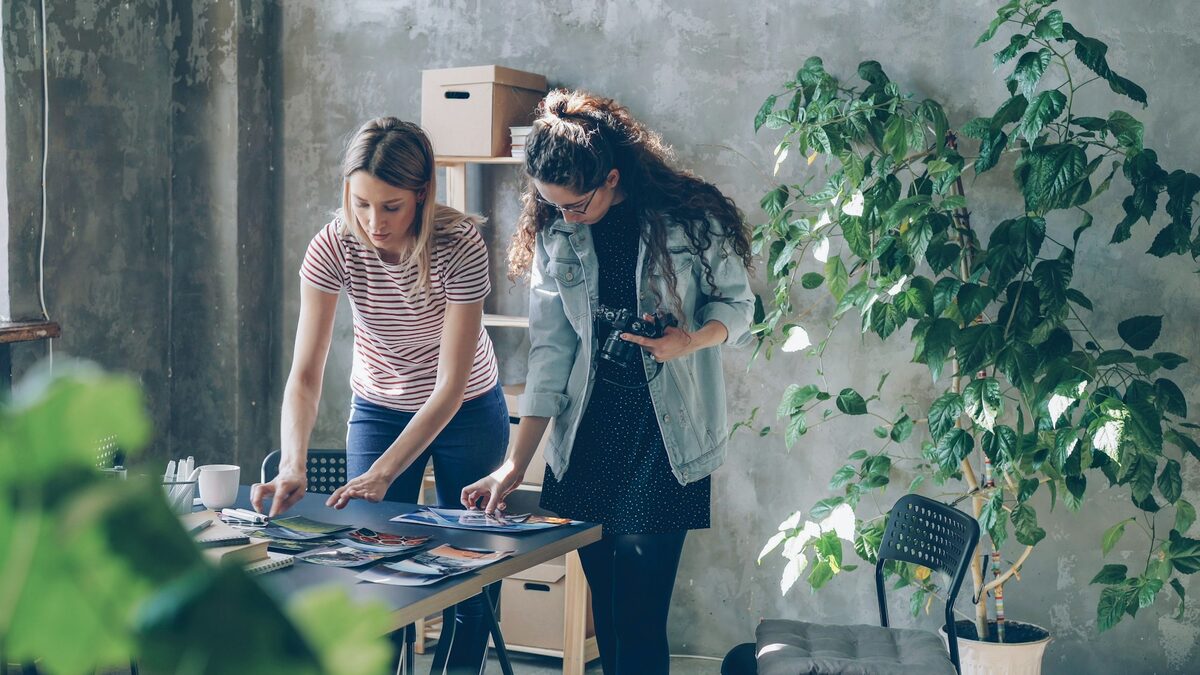 Două colege care aleg fotografii pe masa de lucru, ca să decidă când folosesc imagini pentru firmă create cu AI